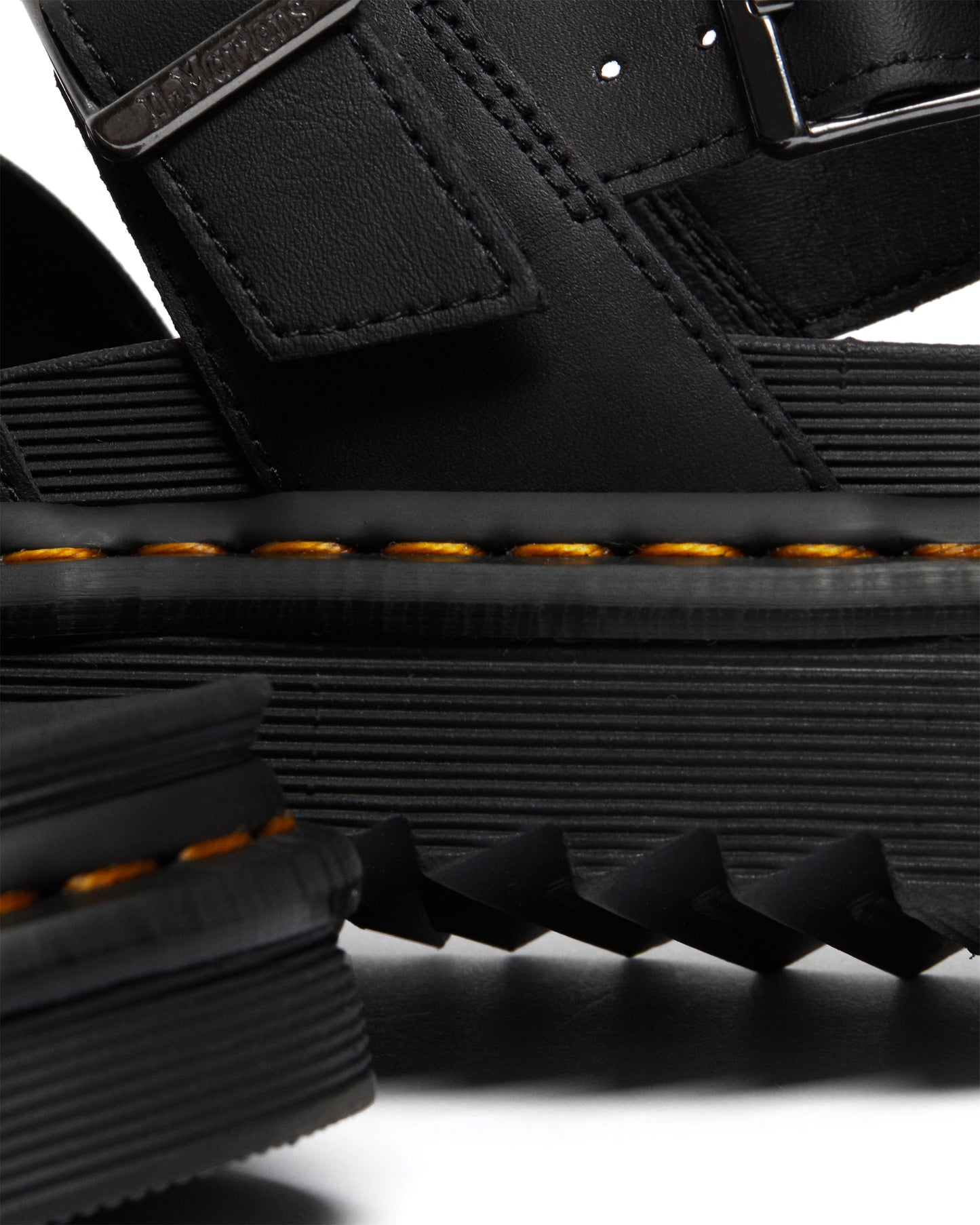 Close-up of black leather sandal featuring buckle strap and thick, ridged sole with yellow stitching.