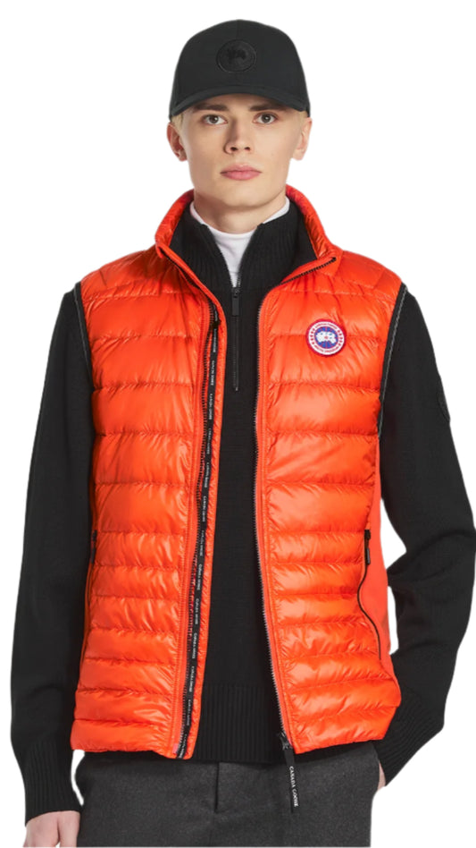 A person wearing an orange puffer vest with the logo on the left chest, over a black sweater and cap.