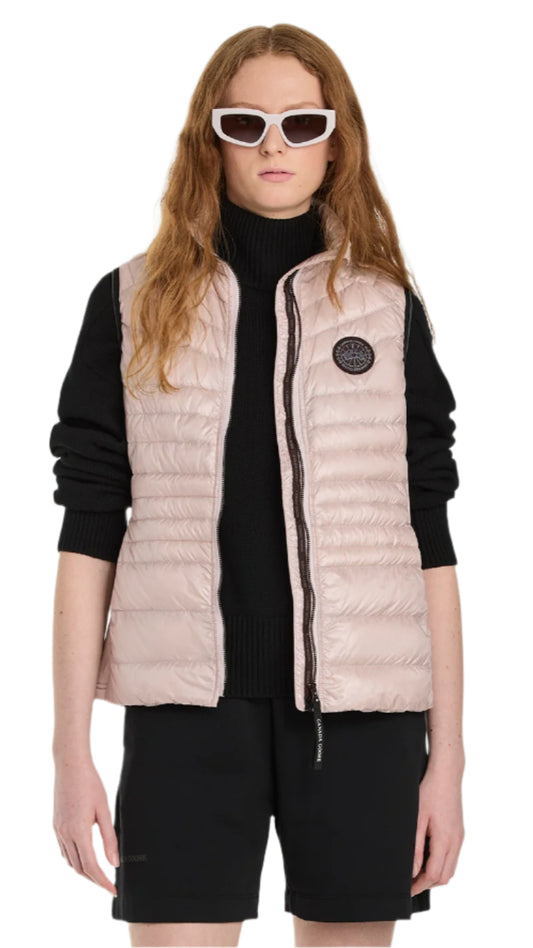A woman in black shorts and a black long-sleeved turtleneck is wearing a light pink, down-filled vest with a full zipper.
