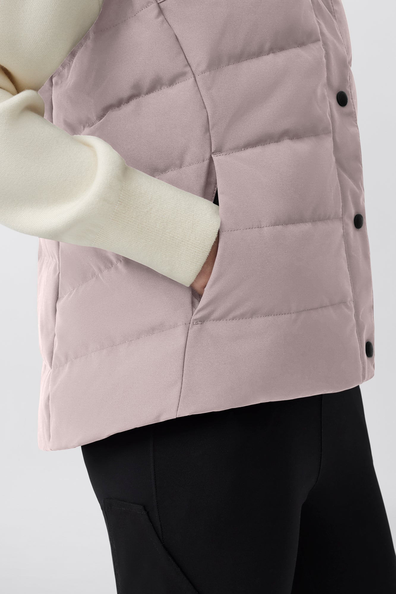 Close-up of a quilted vest in light pink with black snap closures and a hand in a pocket. 