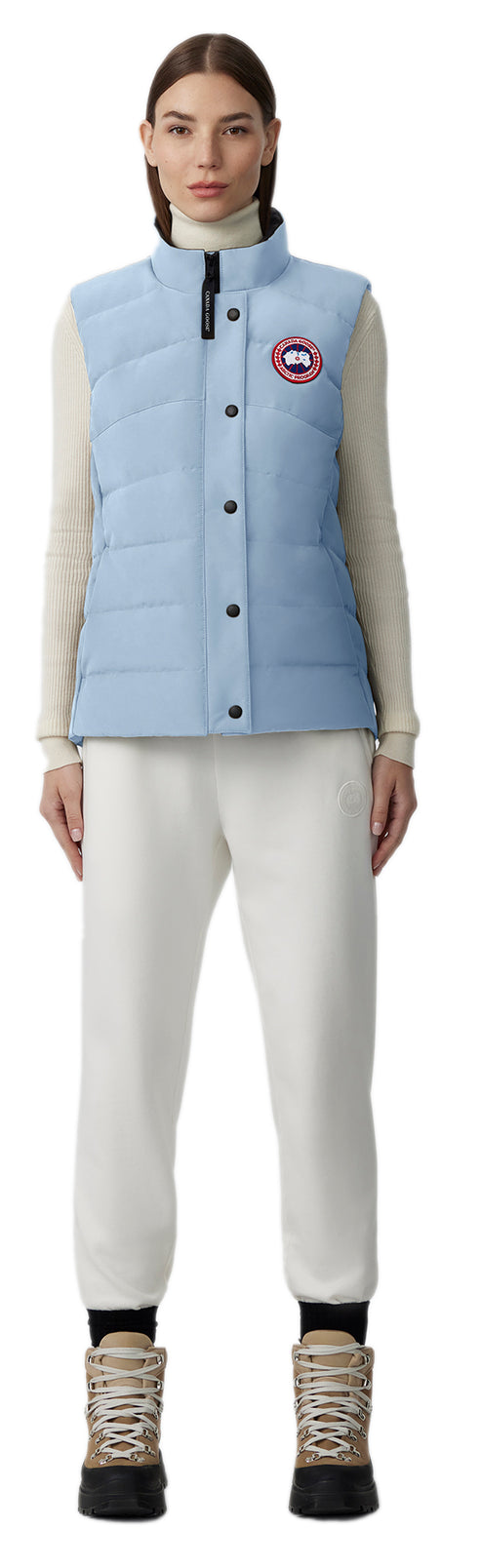A woman wearing a light blue down vest, off-white turtleneck sweater, off-white sweatpants, and black hiking boots.