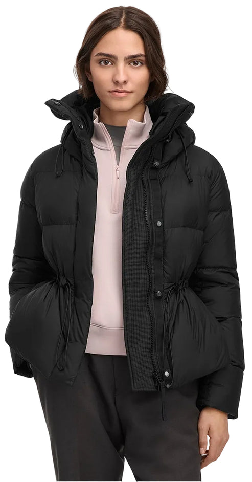 A woman wearing a black puffer jacket with a hood, a cinch-waist drawstring, a stand collar, and a zip-front closure layered over a light pink quarter-zip top and dark pants.