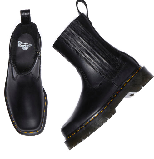 Two black, ankle-high boots with yellow stitching around the soles. One boot is sideways, showing a square toe, side elastic panel, and ridged vamp. The other boot has a zipper, and its interior shows a logo.