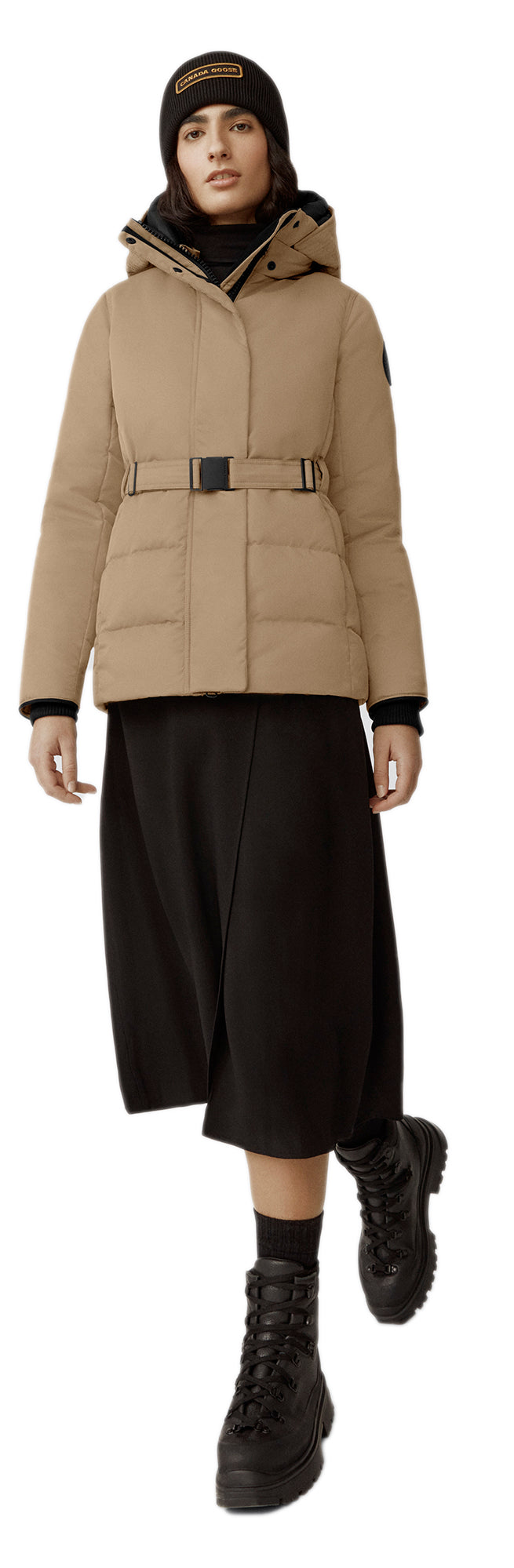 A woman wearing a tan coat with a belt, a black skirt, a black beanie and black boots.