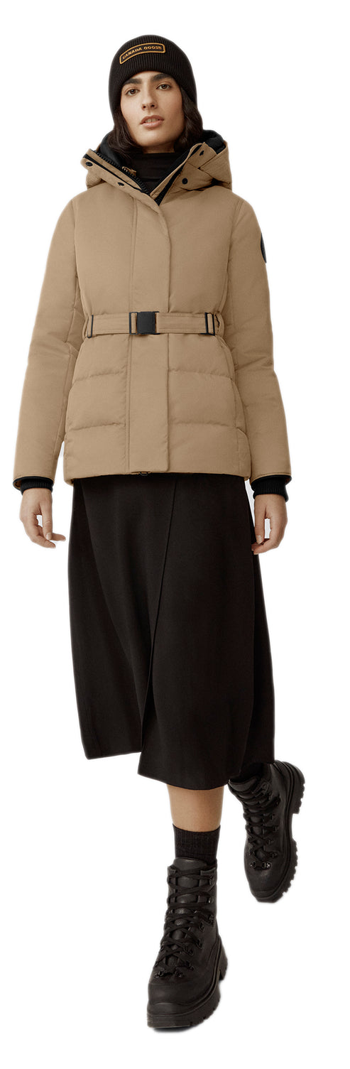 A woman wearing a tan coat with a belt, a black skirt, a black beanie and black boots.