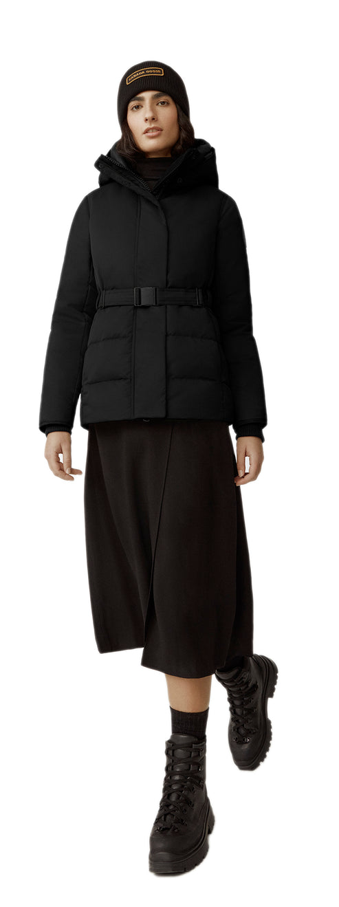 A person wearing a black winter jacket with a belt, a black skirt, black boots and a brown beanie.