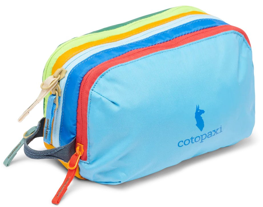 A colorful nylon travel pouch with a light blue front panel featuring a blue llama logo and the text cotopaxi, followed by multi-colored zippered sections in red, blue, yellow, and green, with several zipper pulls and a small side handle.