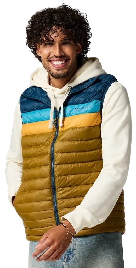 A man with curly dark hair and a beard wearing a white hooded sweatshirt layered under a color-blocked, quilted zip-up vest featuring stripes of dark blue, light blue, yellow, and olive green.