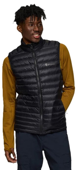 A young man wearing a black down-filled puffer vest over a long-sleeved mustard yellow shirt and navy blue pants, looking slightly to his left.