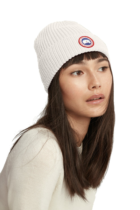 A woman with dark hair wearing a white ribbed beanie with a circular logo.