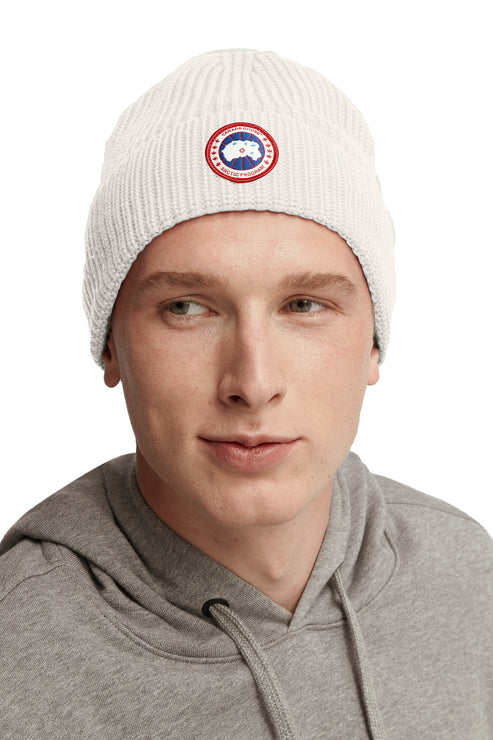 A person wearing a white knit beanie with a round emblem.