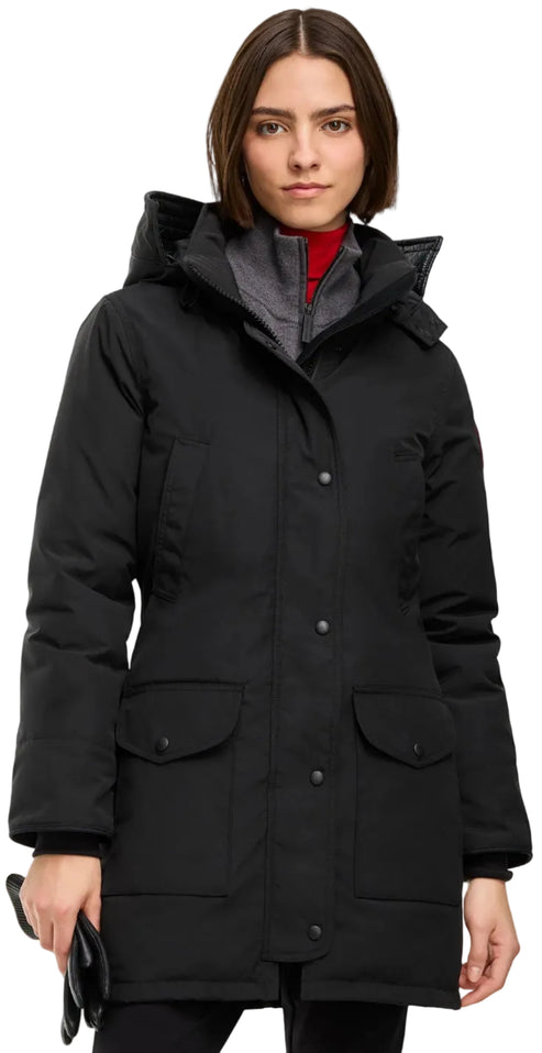 A woman wearing a black, knee-length, down-filled winter parka with a hood, featuring a snap-button front closure, large flap pockets on the front, and a red turtleneck shirt underneath, holding a pair of black gloves.