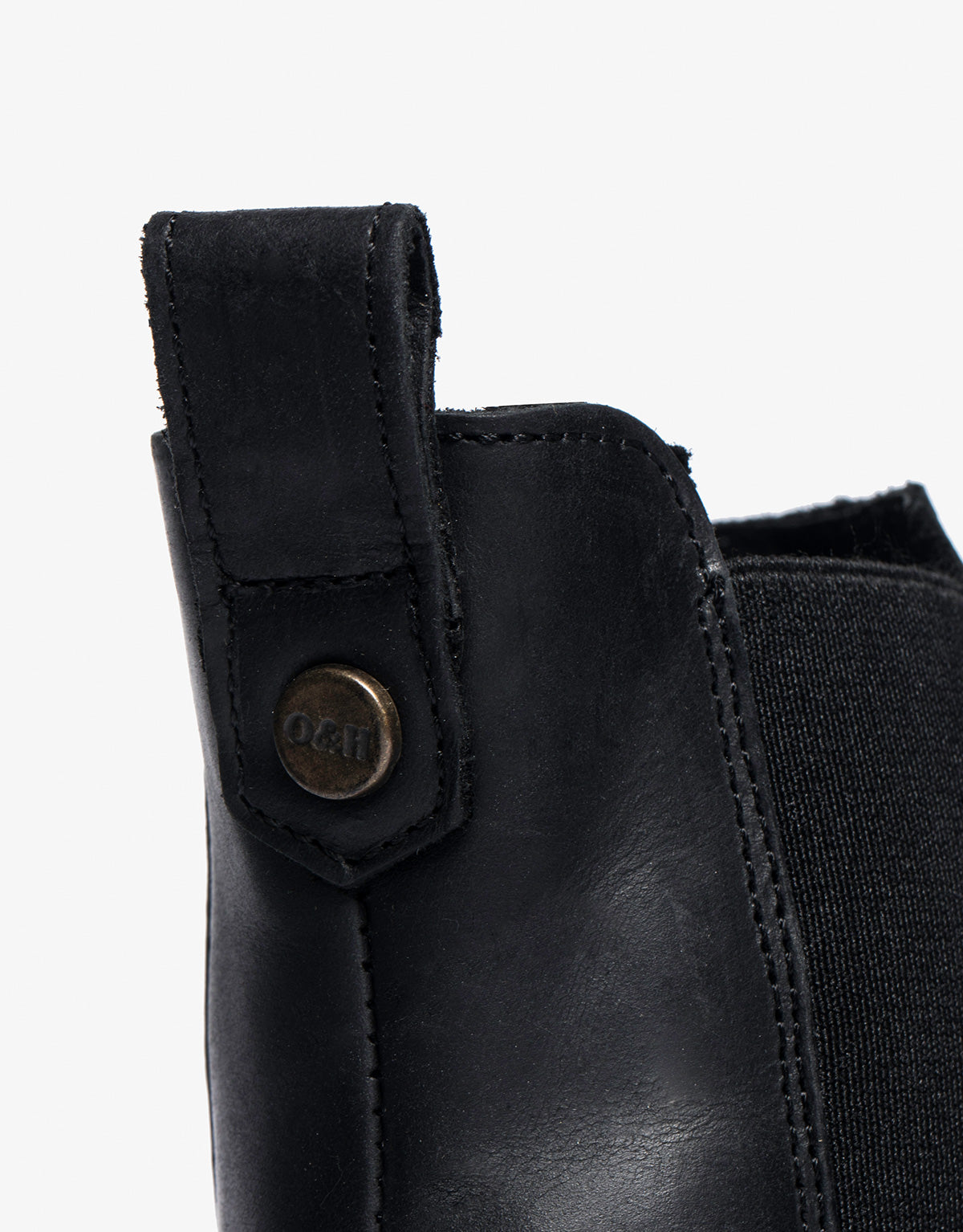 Close-up of a black leather boot showing a heel pull tab with a metal button, which has the letters O&T on it.