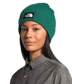 A woman wearing a green beanie with a black square label that has white text.