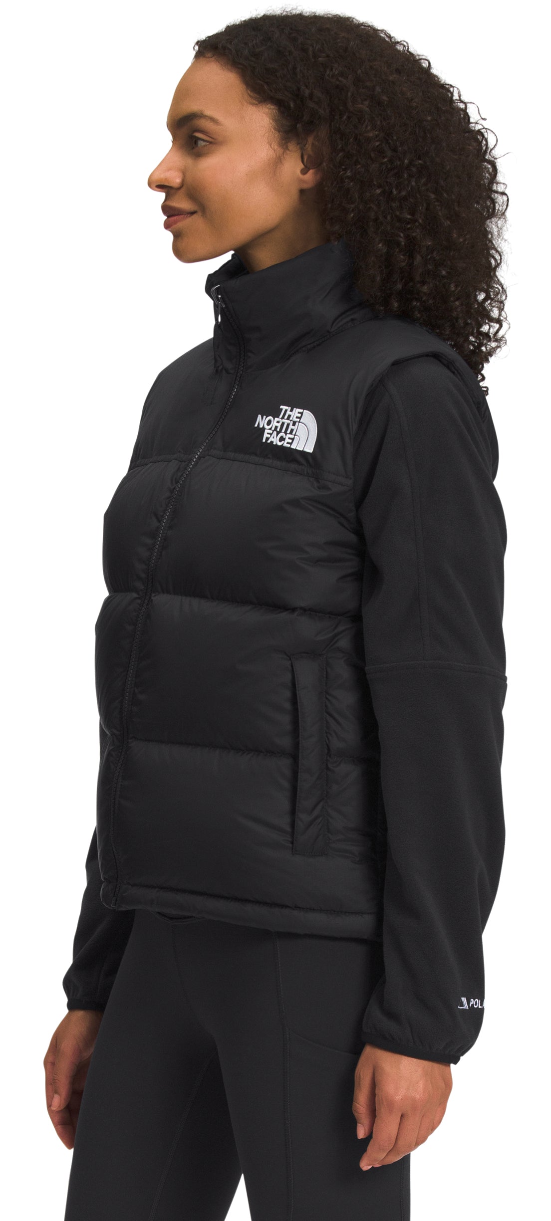 A woman wears a black puffer vest with the brand's logo on the left chest over a long-sleeve black shirt and leggings.
