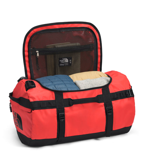An open red duffel bag with black straps and bottom, revealing folded clothing and a mesh compartment with a logo.