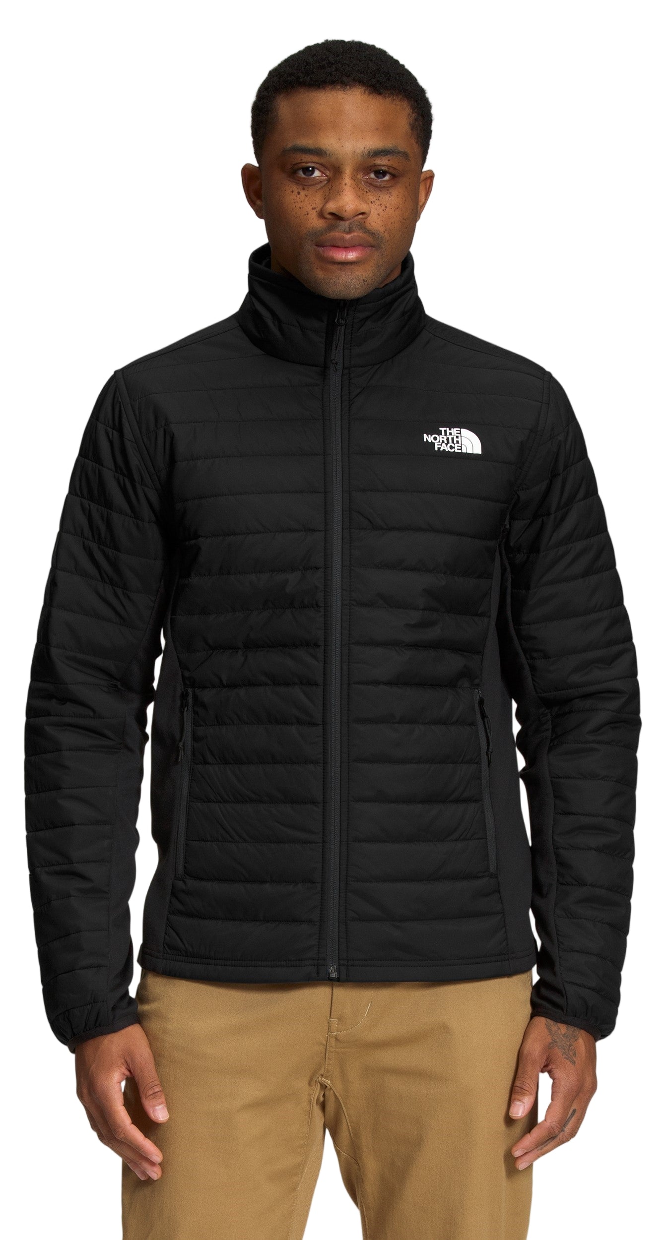 A man wears a black padded jacket with a white logo on the left chest, and tan pants.