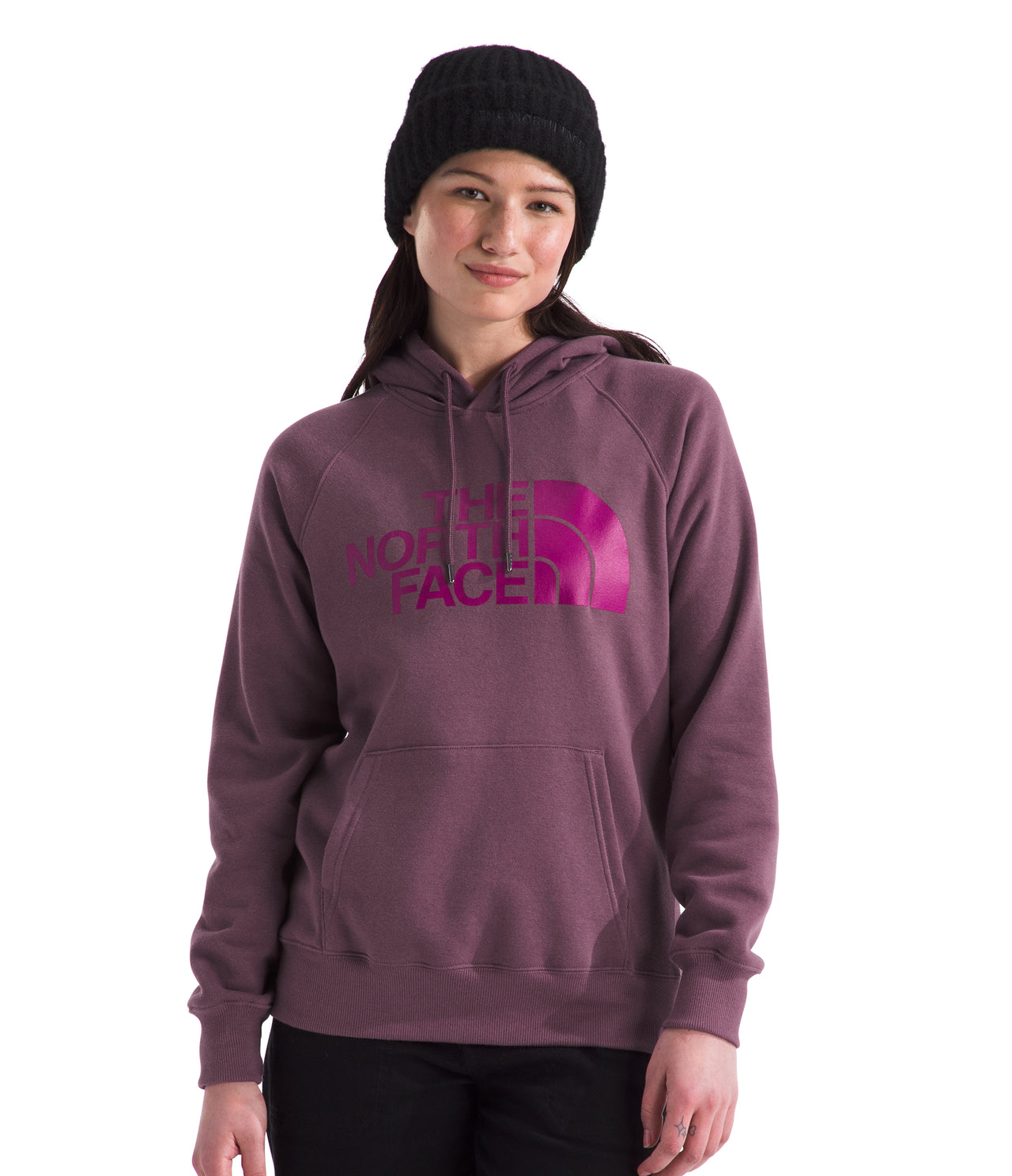 A woman in a black beanie and a plum-colored hooded sweatshirt with magenta text on the front is standing.
