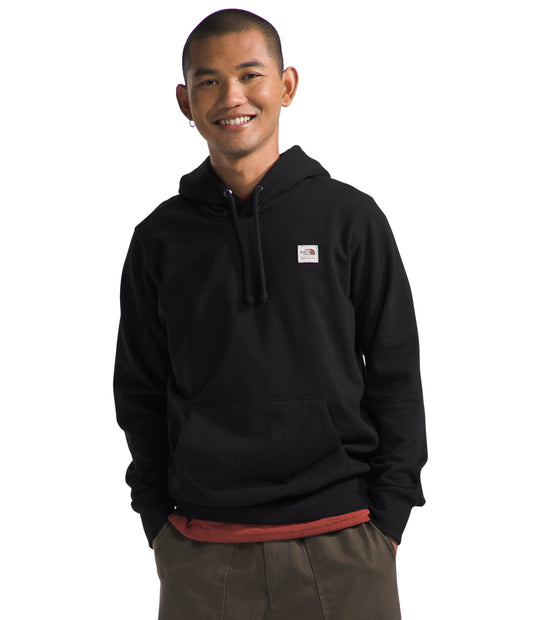 A smiling person wearing a black hooded sweatshirt with a logo patch on the chest.