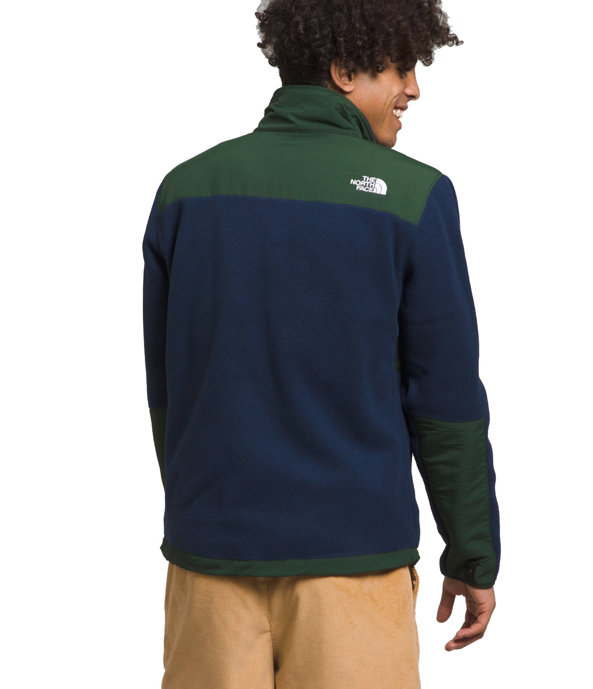 Man wearing a color-blocked fleece jacket in navy, forest green, and tan pants. The jacket has a high collar and a small logo on the upper back.