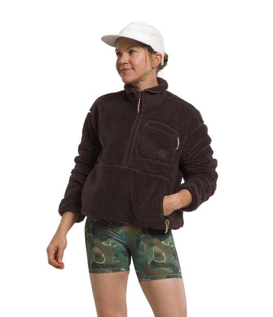 A person wearing a dark brown, long-sleeve fleece jacket with a pocket and a white cap, paired with green and brown camo shorts.