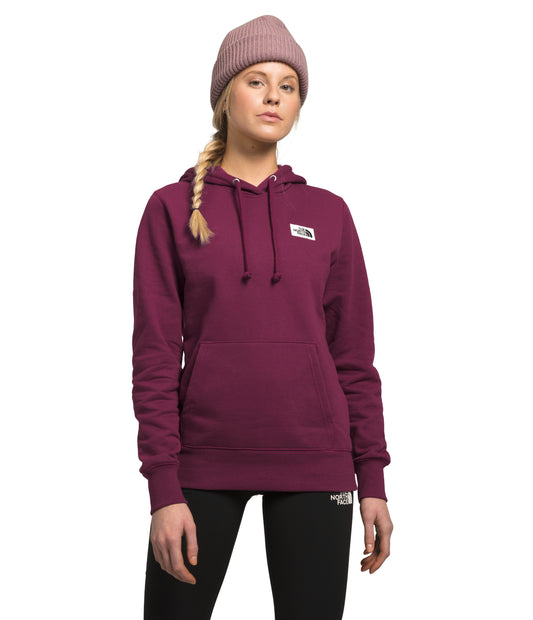 A woman wearing a dusty pink beanie and a dark burgundy hoodie with a square label on the chest.