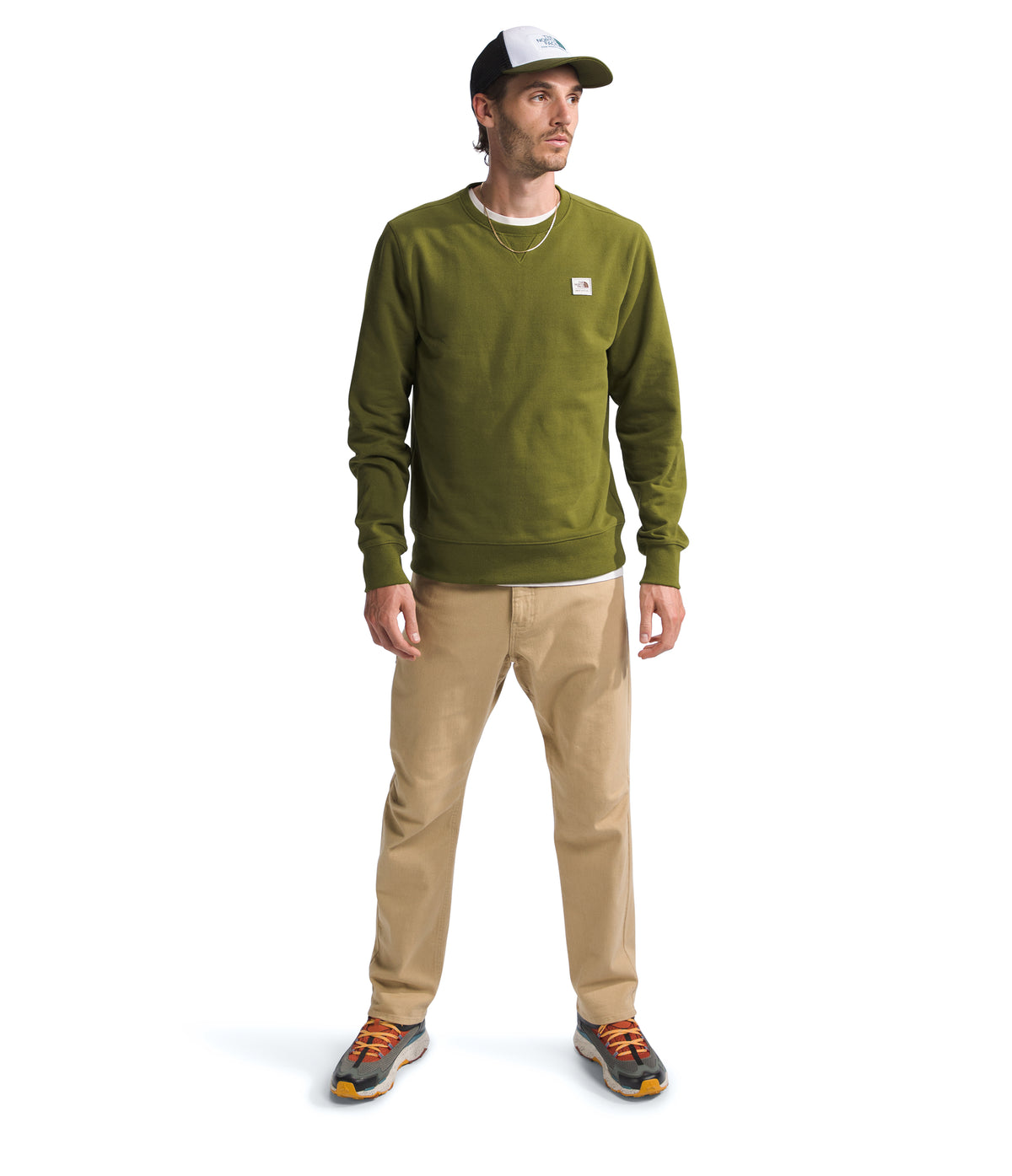 A man wearing a dark green crew neck sweater, khaki pants, and shoes and a trucker hat, posing.