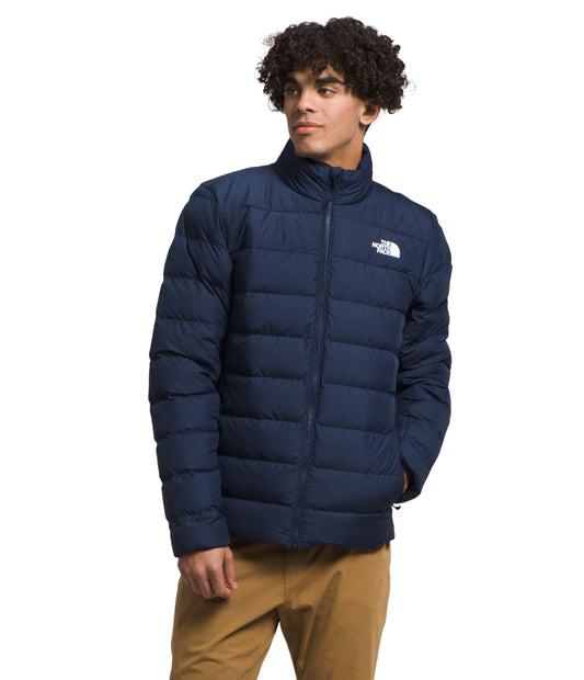 A male model wears a quilted, navy-blue puffer jacket and khakis. The jacket is zipped up and has a small, white logo on the upper right chest.