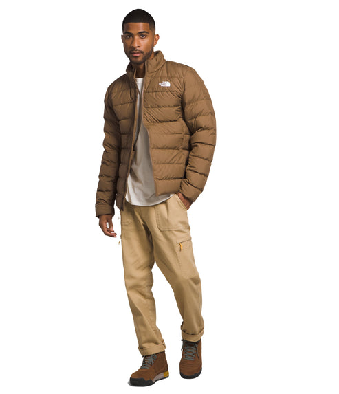 A male model wears a brown puffer jacket, a white shirt, tan pants, and brown hiking boots.