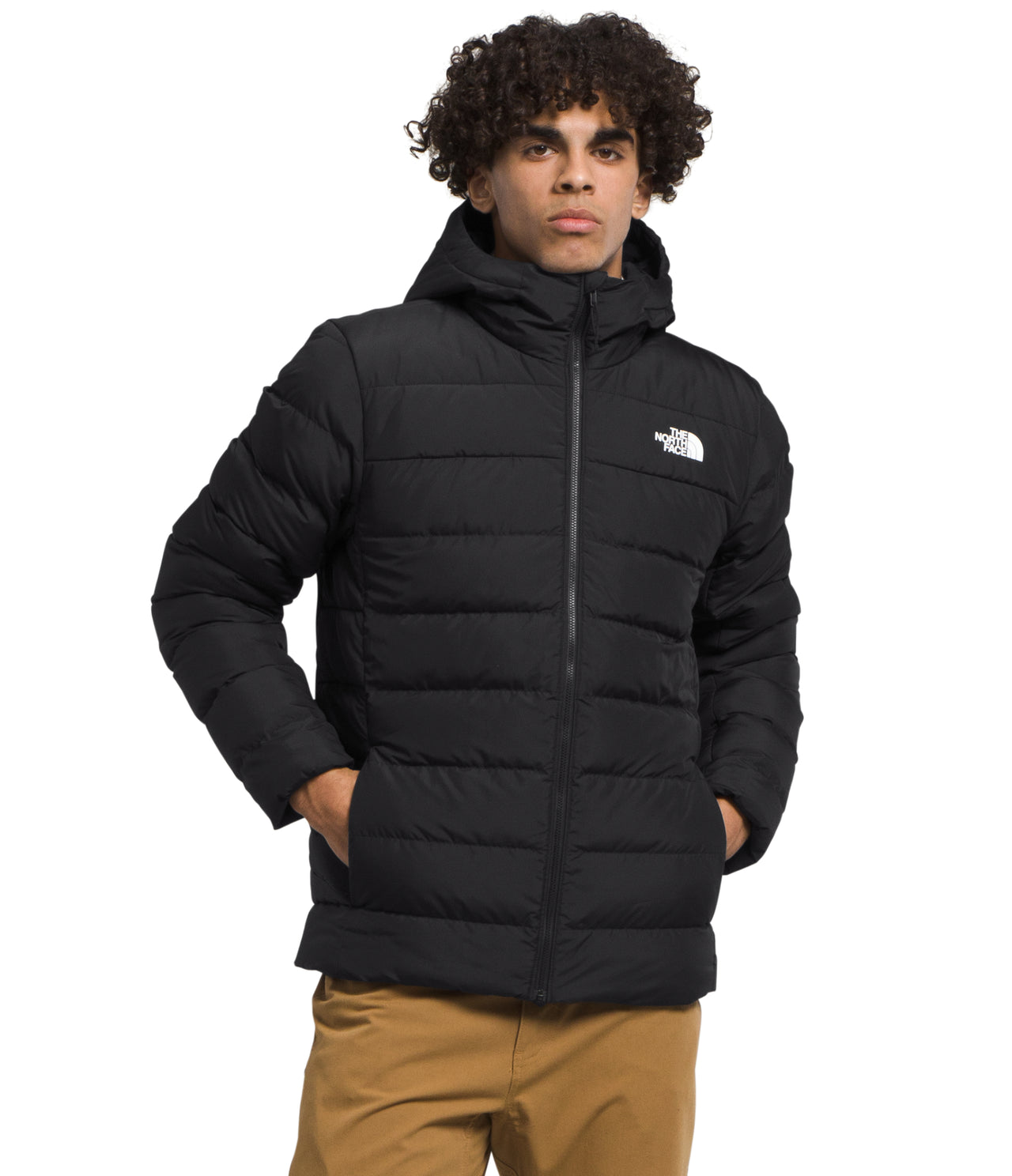 A man is standing wearing a black hooded puffer jacket with his hands in his pockets.