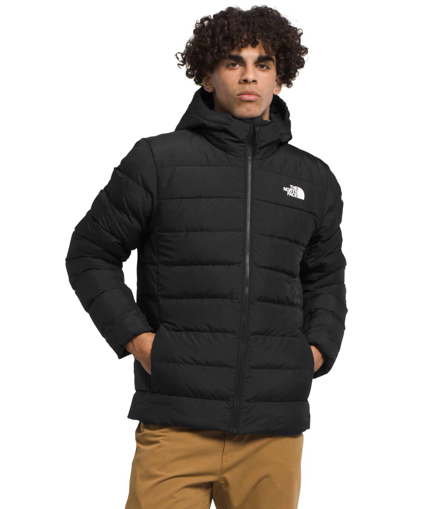 A man is standing wearing a black hooded puffer jacket with his hands in his pockets.