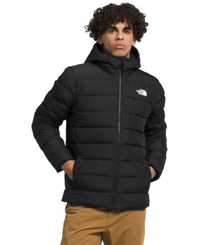 A man is standing wearing a black hooded puffer jacket with his hands in his pockets.