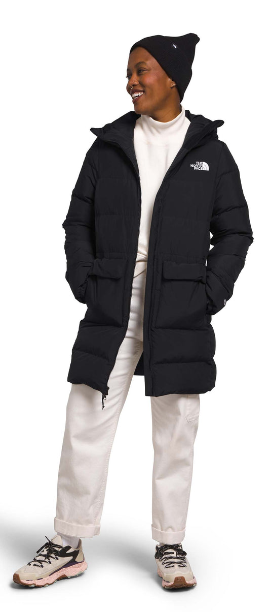 A person wears a black, long puffer coat with a hood and a black beanie with white pants and sneakers.