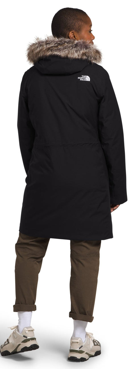 A person wearing a black parka with a fur-lined hood, brown pants and white socks and sneakers.