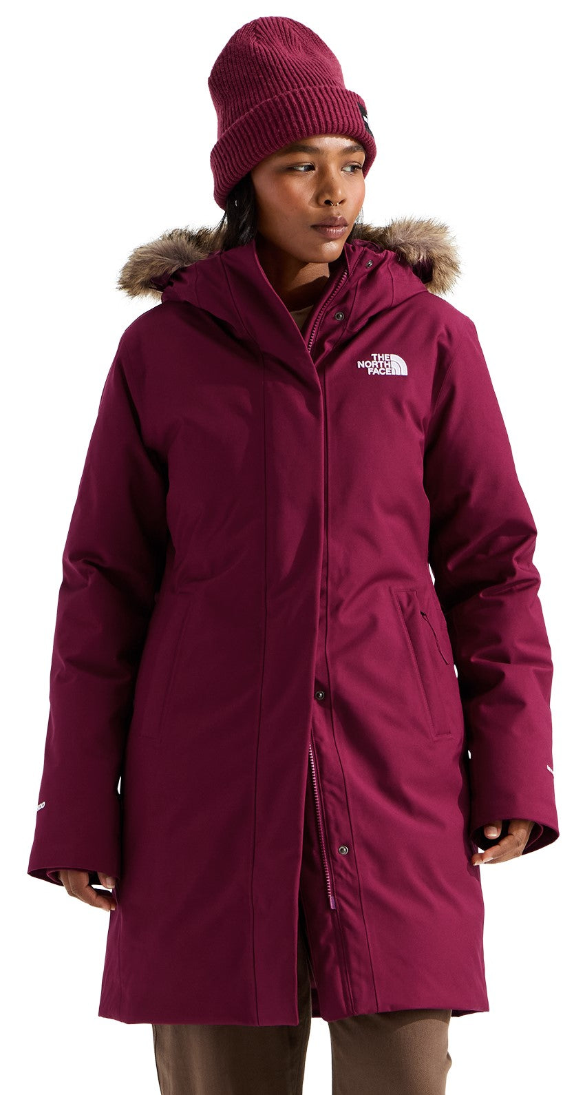 A woman wearing a knee-length, deep magenta, insulated parka with a faux-fur-trimmed hood and a matching beanie.