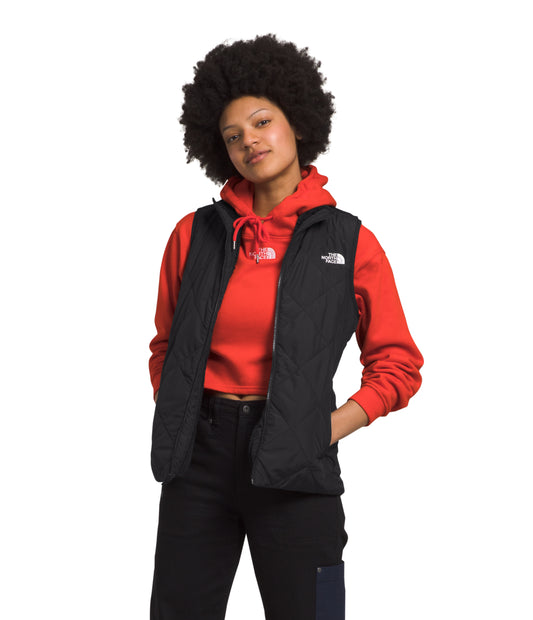 A woman with dark, curly hair stands with her hands in her pockets while wearing a black vest over a red hooded top and dark jeans.