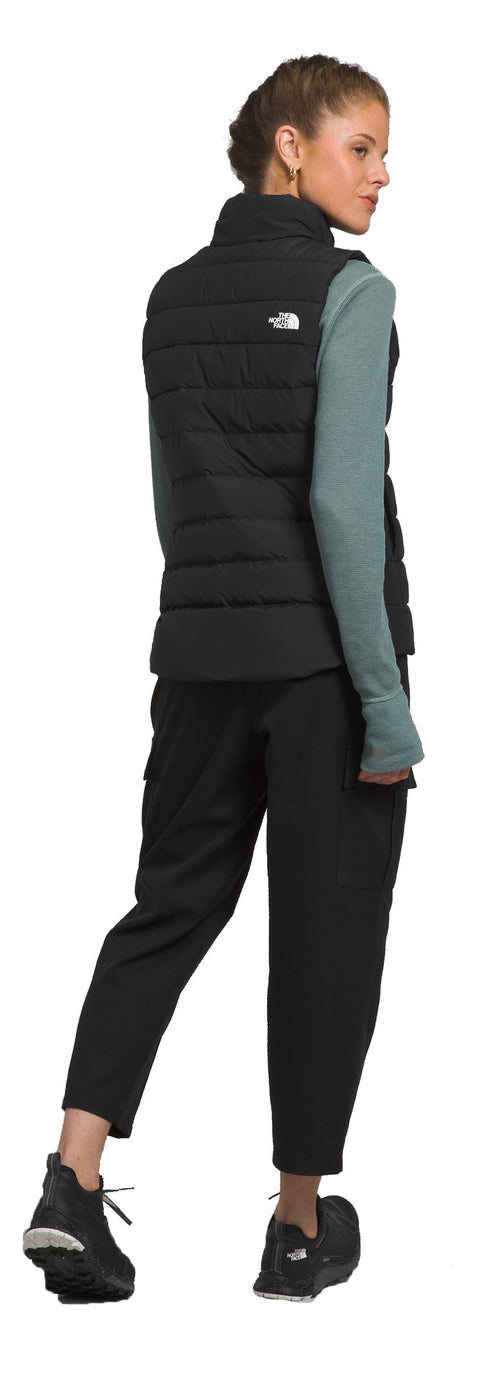 A woman wearing a black vest, bluish green long sleeve shirt, black pants, and black shoes looks to the side.