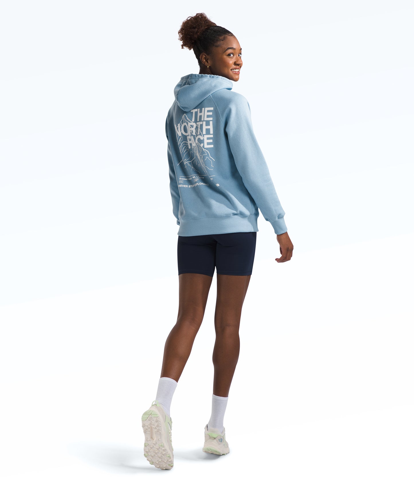 A woman wearing a light blue hoodie with "THE NORTH FACE" on the back and navy shorts, looking over her shoulder.