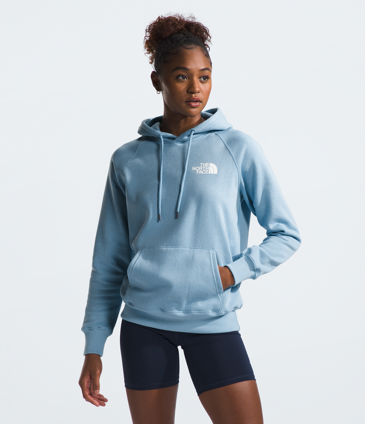 Woman in light blue hoodie with white logo and dark shorts, standing.