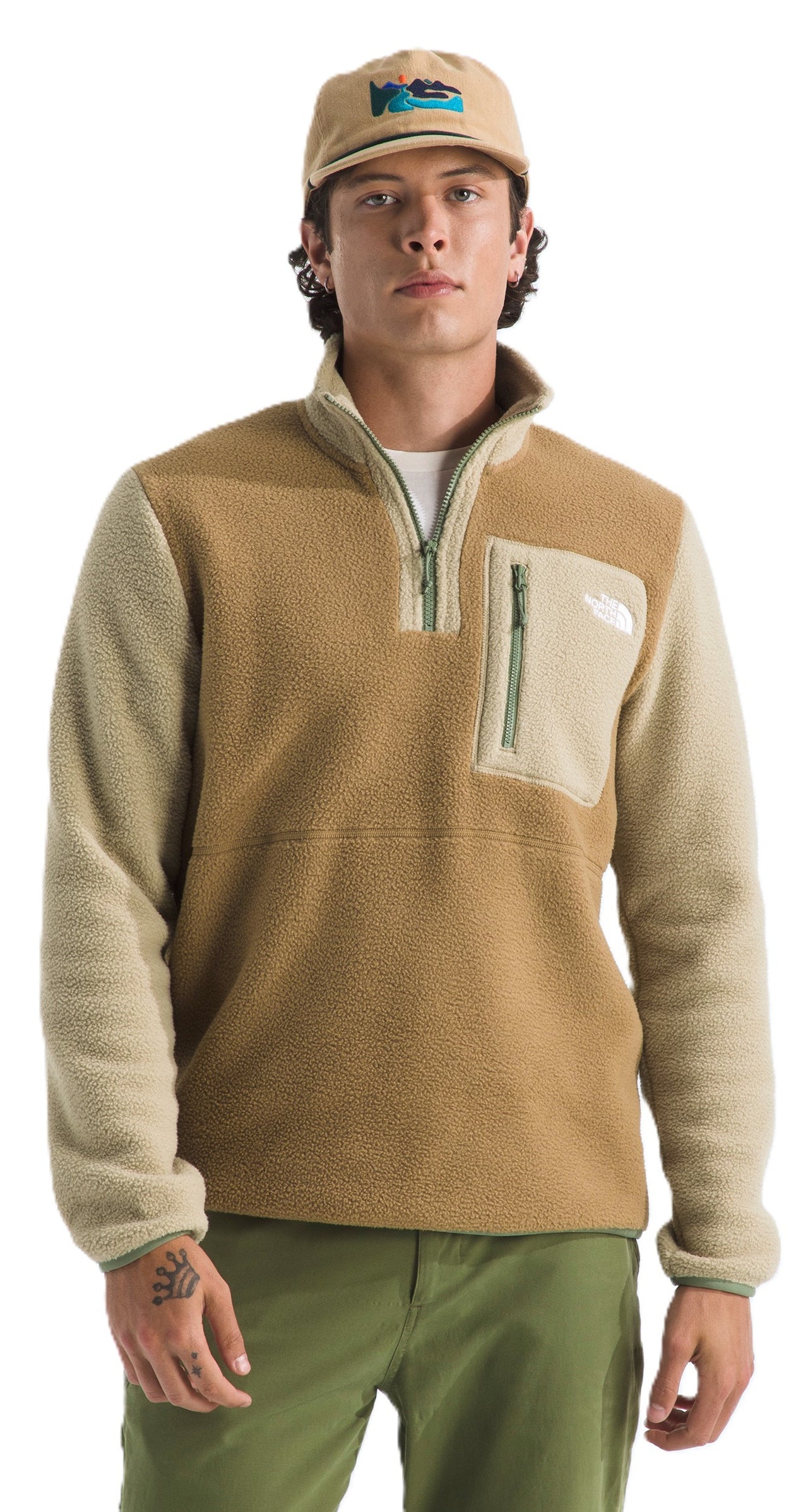 A person wearing a tan, half-zip fleece with tan sleeves, a front pocket with a zipper, green pants, and a matching hat.