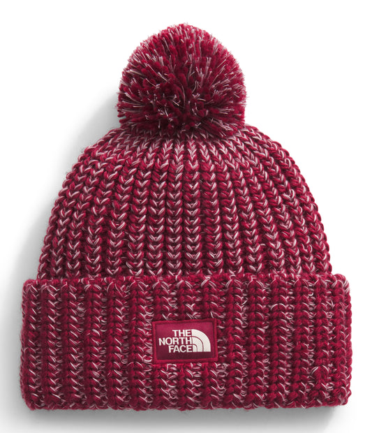 Close-up of a burgundy ribbed knit beanie with a pom-pom on top and a rectangular white label with text on the front.