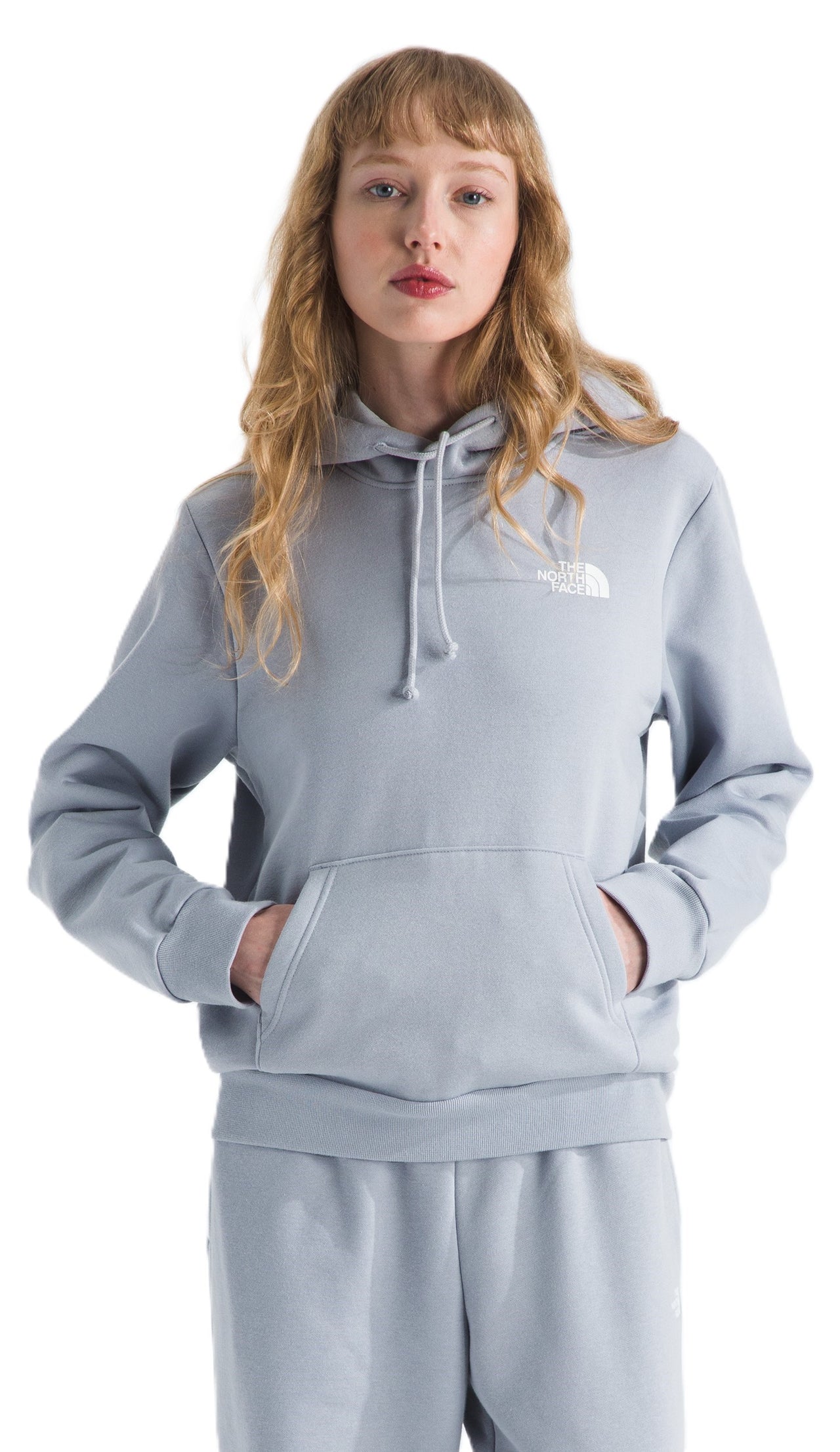A person stands wearing a pale blue hooded sweatshirt with white logo and a kangaroo pocket.