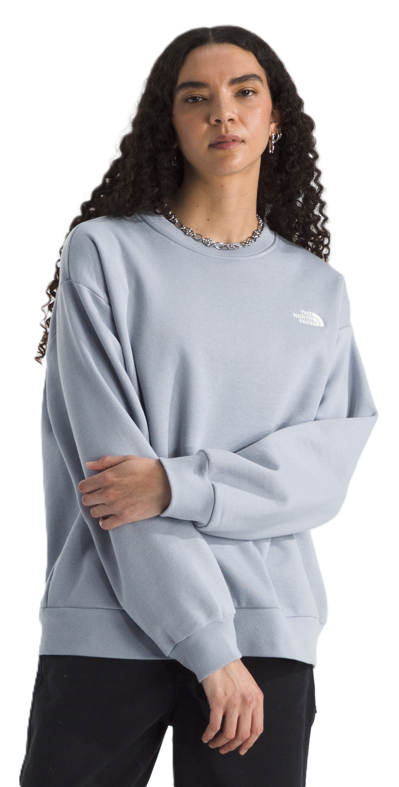 A woman wearing a light blue, long-sleeved sweatshirt with a small logo on the upper left chest, and a silver chain necklace.