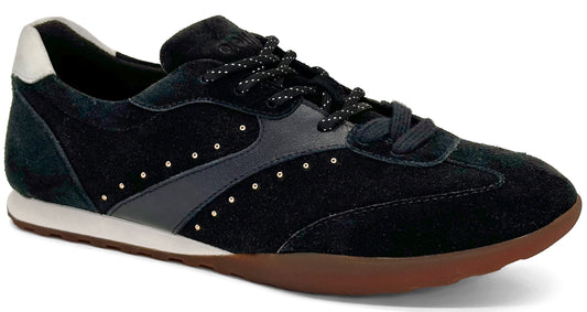 A black suede sneaker with a white heel tab, white midsole, and gum rubber outsole, featuring a dark grey side panel adorned with small metallic studs and black patterned laces.