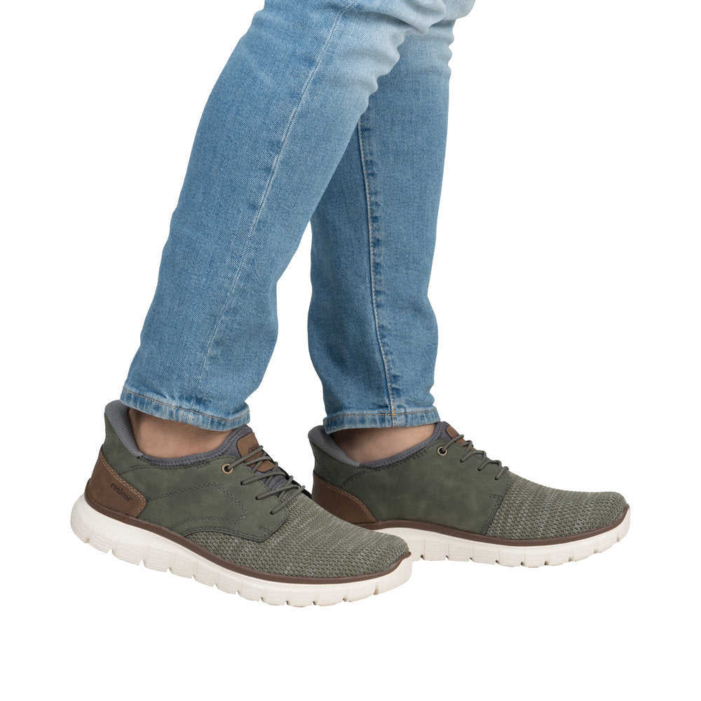 A person wearing olive green sneakers with light blue jeans. The sneakers have brown leather accents and white soles.
