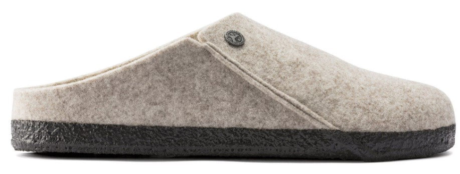 Side view of a beige felt slip-on slipper with a dark brown textured rubber sole and a small metallic circular emblem on the side.