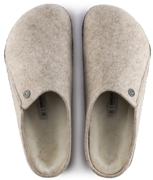 A pair of beige felt slip-on clogs lined with white shearling, featuring a small metallic rivet on each side and the text 'BIRKENSTOCK' printed on the insole.