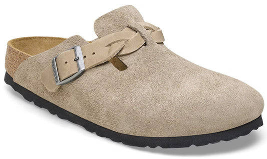 A beige suede clog-style shoe with a braided leather strap and an adjustable metal buckle across the instep, featuring a natural cork footbed and a textured black rubber sole.