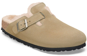 A tan suede slip-on shoe lined with cream-colored faux fur, featuring an adjustable side strap with a metallic buckle and a dark, textured rubber outsole.
