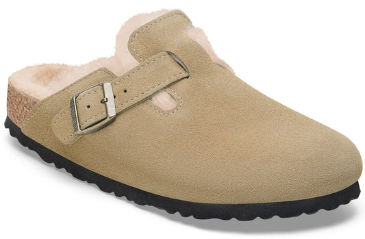A tan suede slip-on shoe lined with cream-colored faux fur, featuring an adjustable side strap with a metallic buckle and a dark, textured rubber outsole.